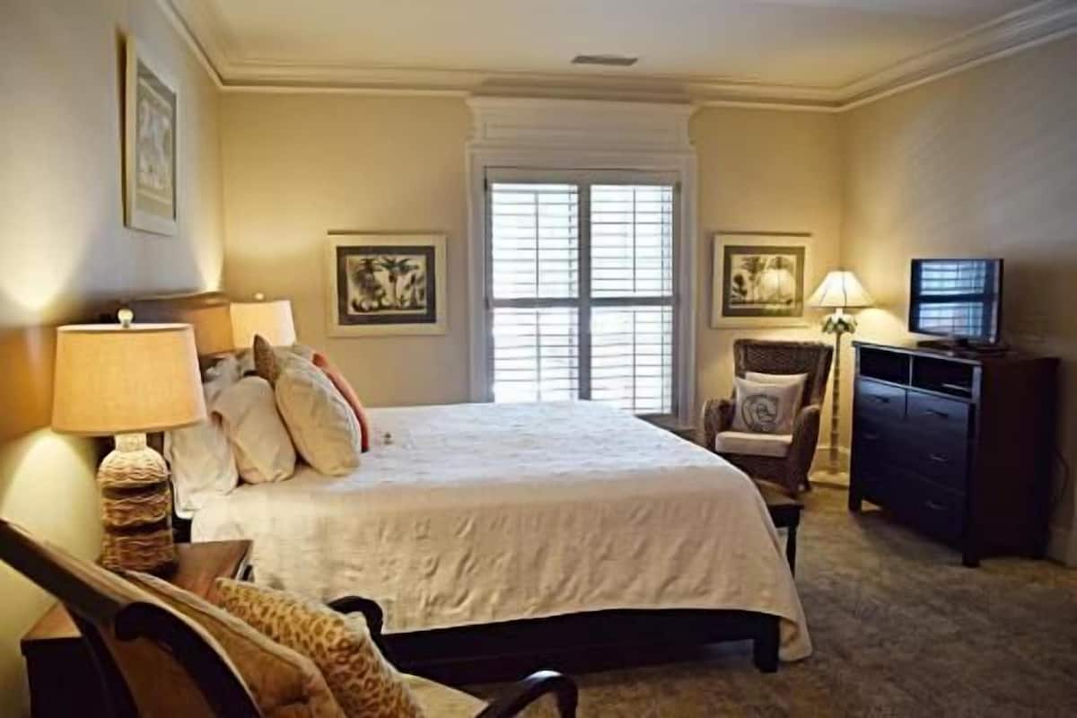 A bedroom with a large bed, a chair, a desk, a television, and a window with blinds.