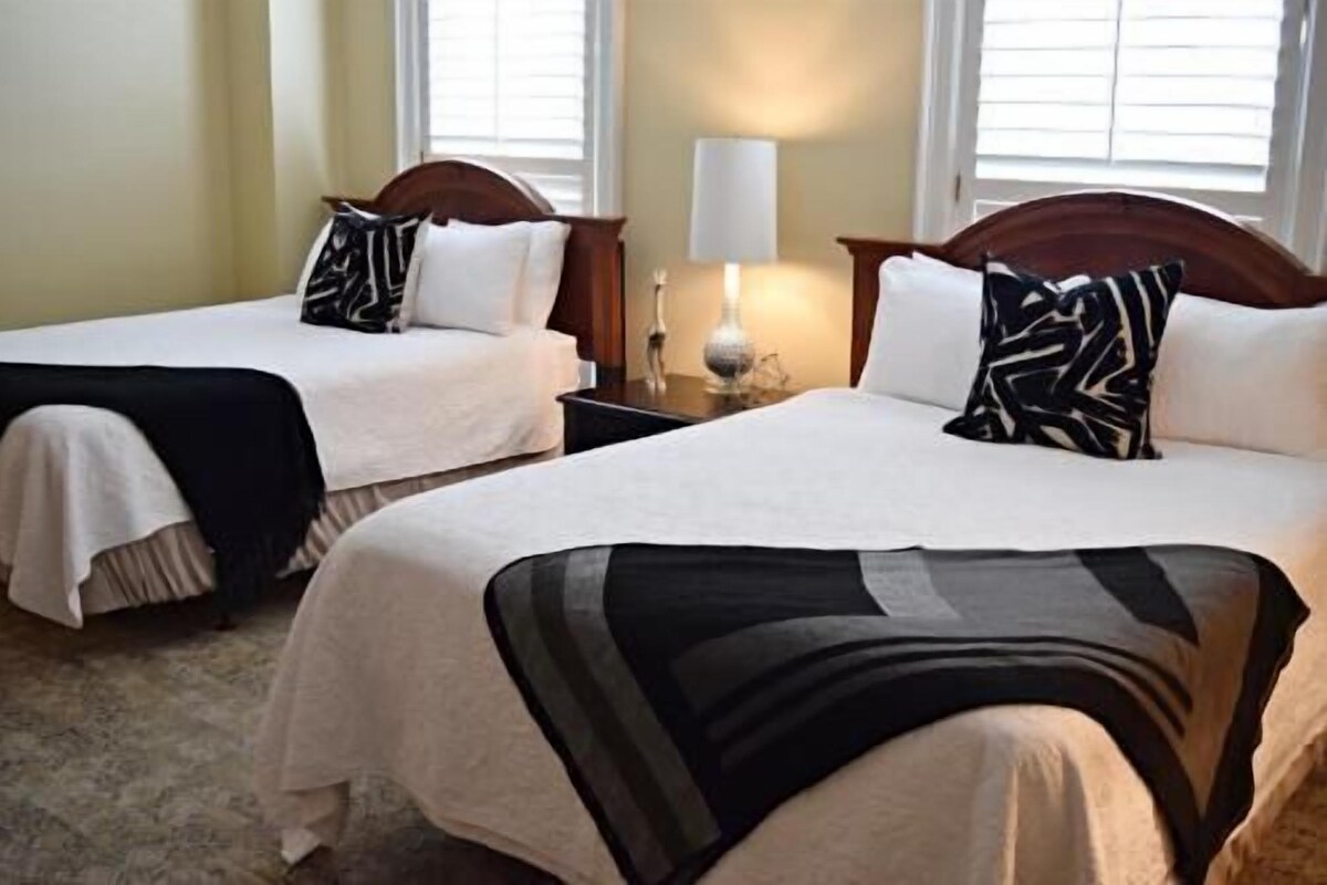 Two adjacent beds with white bedding, a nightstand with a lamp, and a window with blinds.