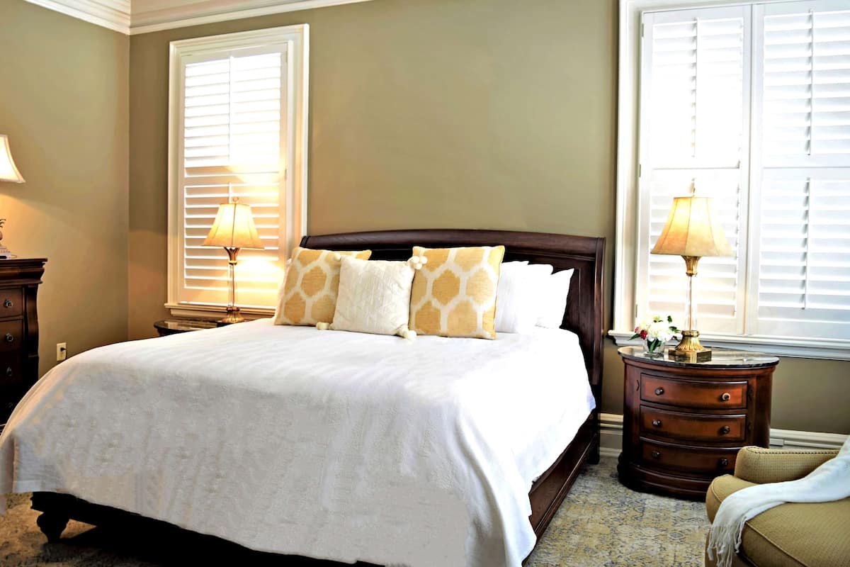 A neatly made bed with a white comforter and patterned pillows, flanked by two bedside tables with lamps, in a room with large windows and a carpeted floor.