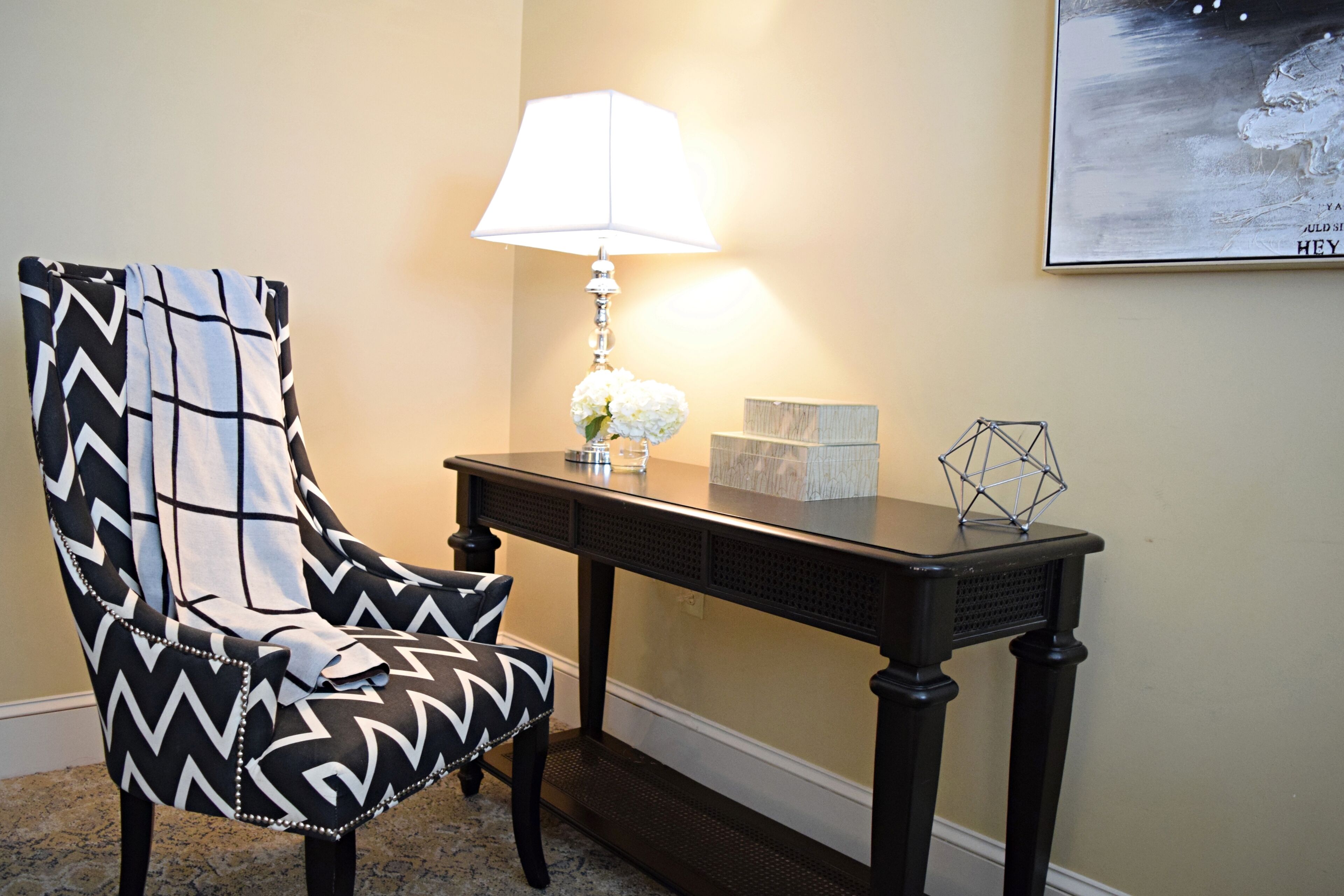 A chair with a black and white zigzag pattern, a table with a lamp, and a framed picture on the wall.
