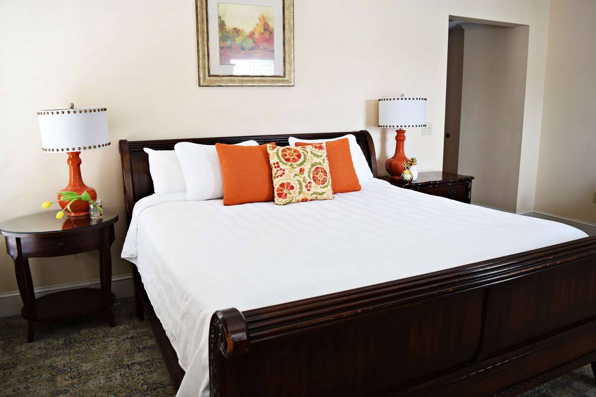 A neatly made bed with white linens and orange and patterned pillows, flanked by two wooden nightstands with lamps, in a room with a framed picture on the wall.