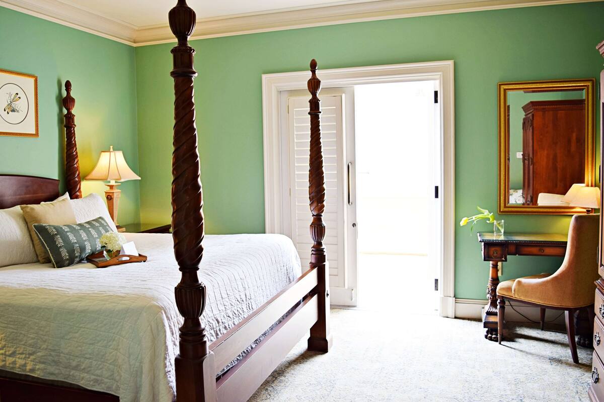 A bedroom with a four-poster bed, a wooden chair, a desk with a lamp, and a mirror on the wall.