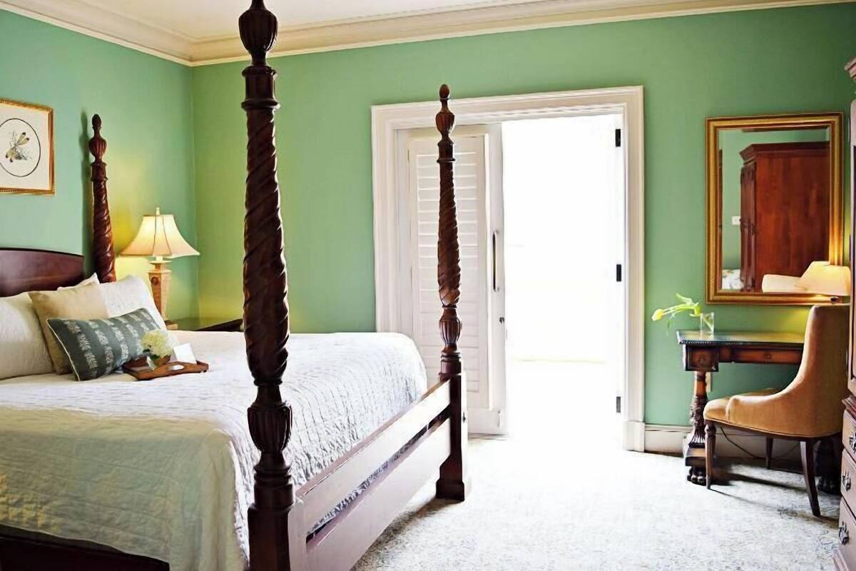 A four-poster bed, a desk with a chair, a mirror, and a door leading to another room.