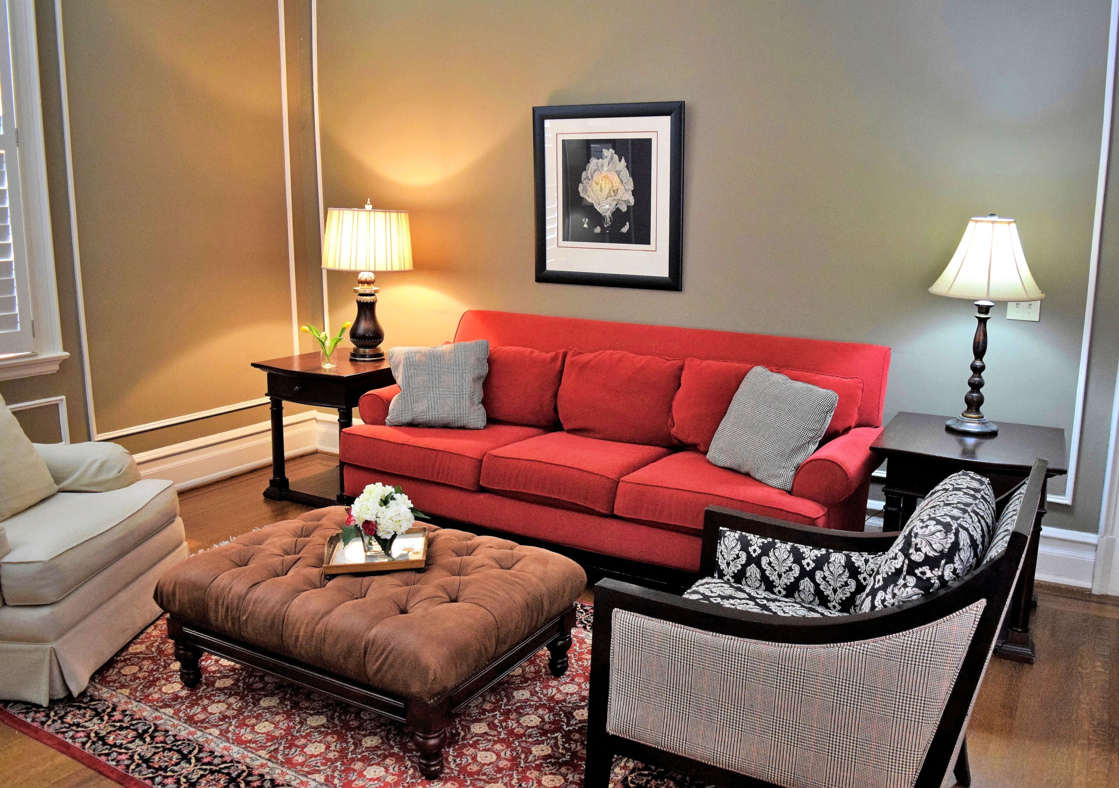 A living room with a red sofa, a brown ottoman, a patterned armchair, and a small table with a lamp.