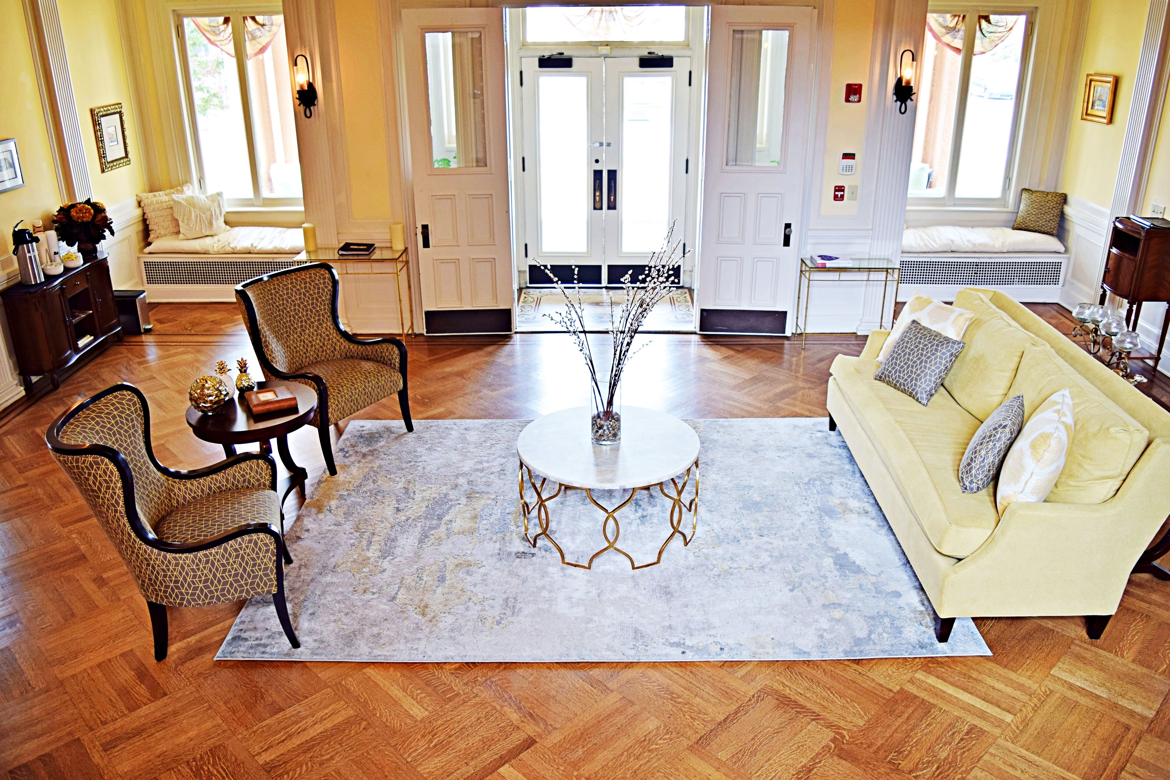 Un salon spacieux avec un canapé jaune, deux fauteuils à motifs, une table basse ronde et une grande fenêtre avec des rideaux.