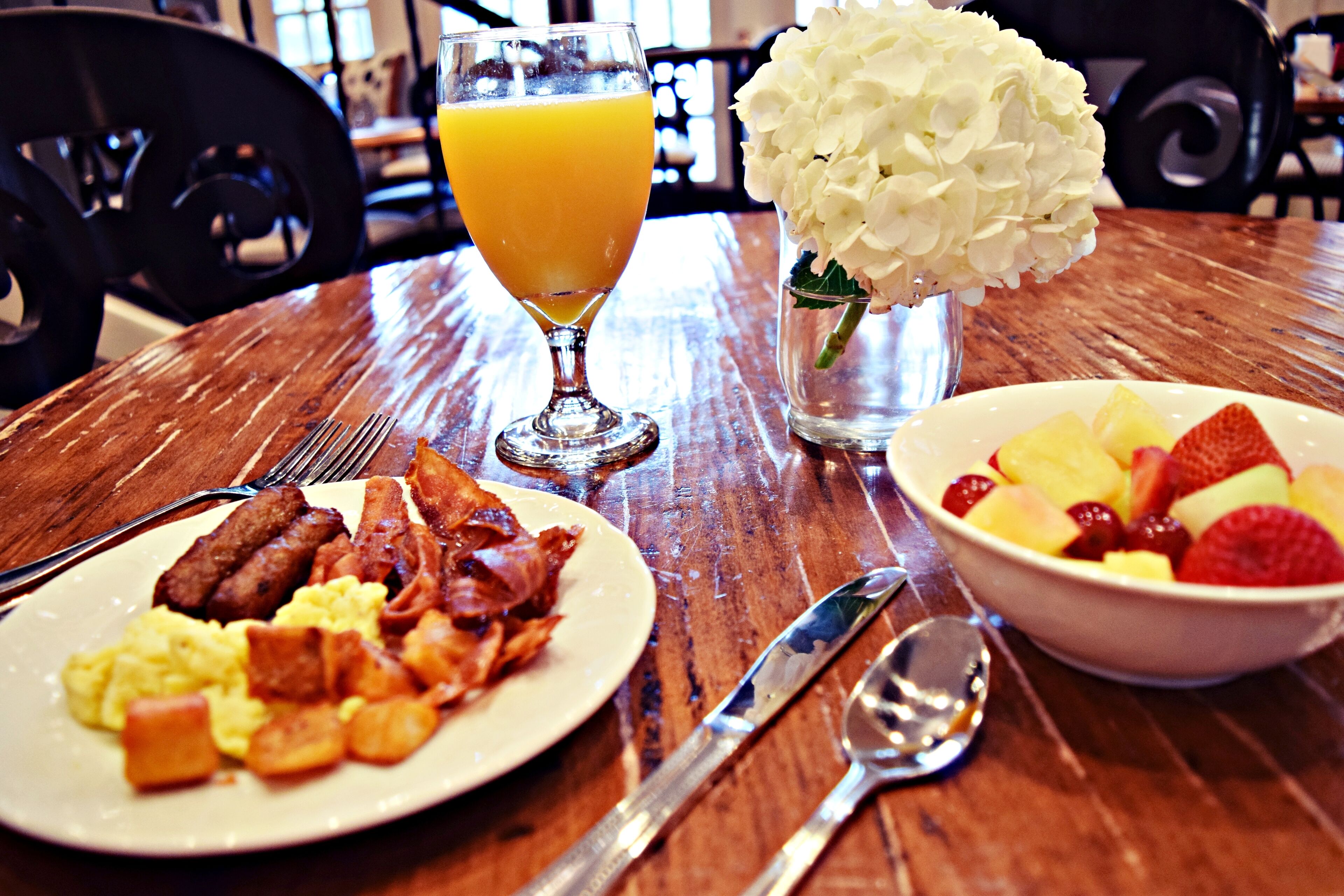 A breakfast setting with a plate of eggs, bacon, and potatoes, a glass of orange juice, a bowl of fruit, and a vase of white flowers on a wooden table.