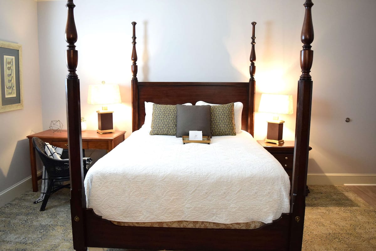 A four-poster bed with a white quilt, two bedside lamps, and a chair with a patterned cushion.