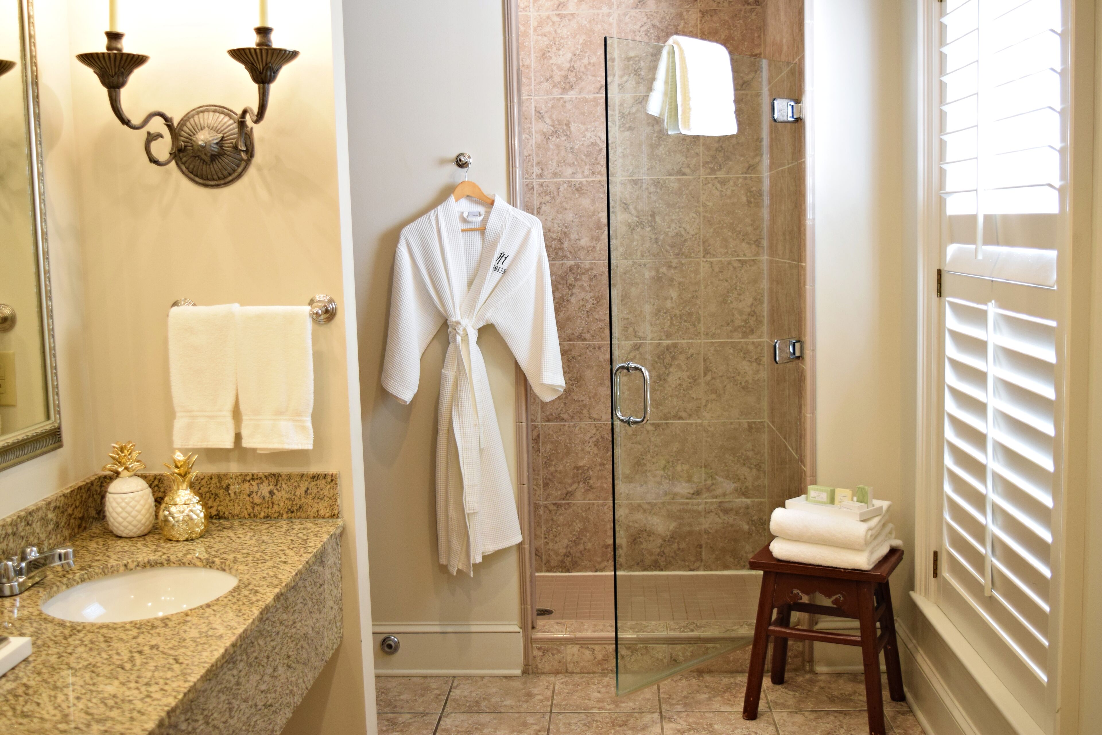 Un baño con lavamanos de encimera de granito, cabina de ducha de vidrio y taburete de madera con toallas.