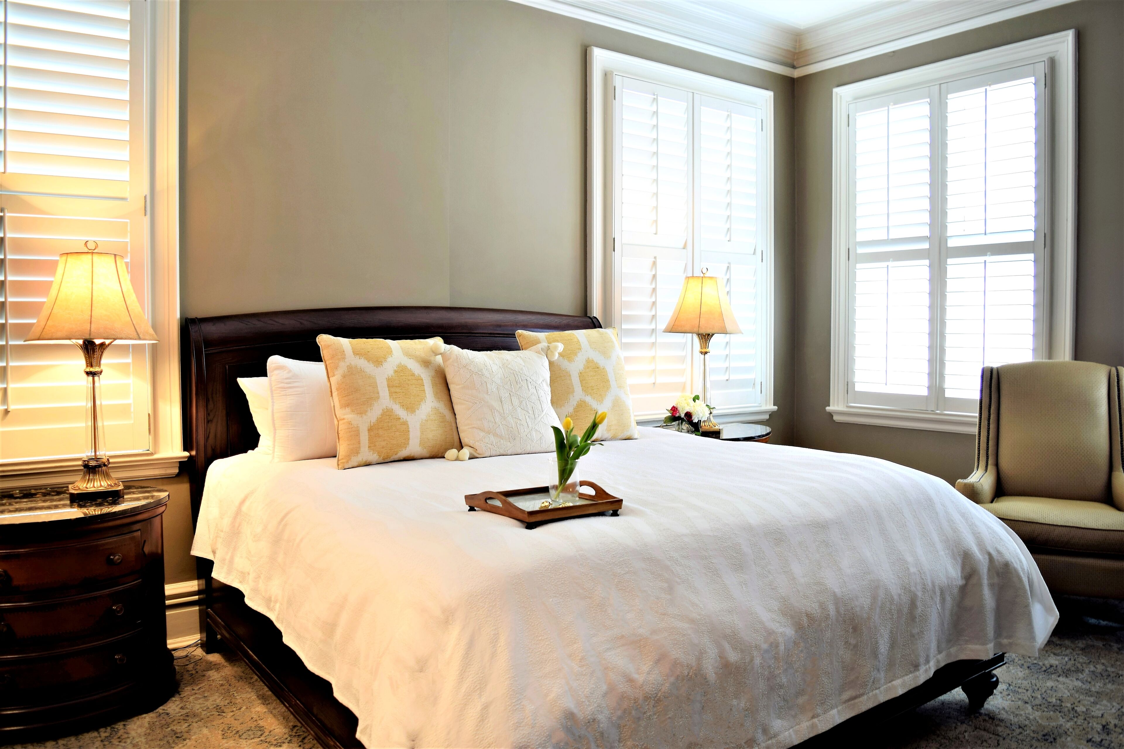 A neatly made bed with white bedding and patterned pillows, flanked by bedside tables with lamps, in a room with large windows and a chair.