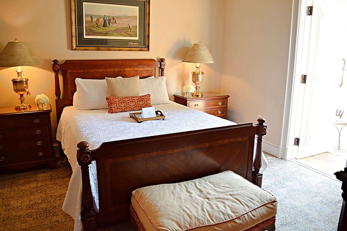 A neatly made bed with a wooden headboard, two bedside tables with lamps, a bench at the foot of the bed, and a framed picture on the wall.