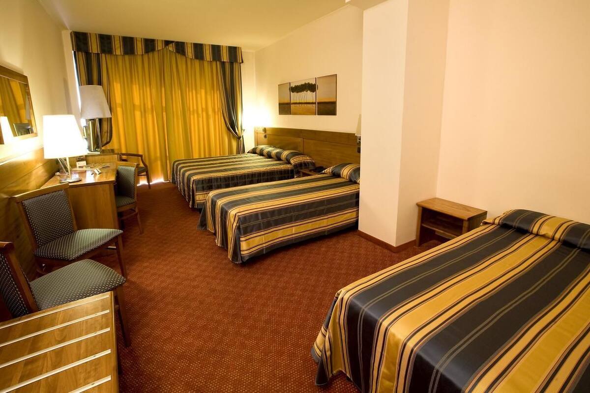 A hotel room with four single beds, a desk with a chair, a lamp, and a small table.