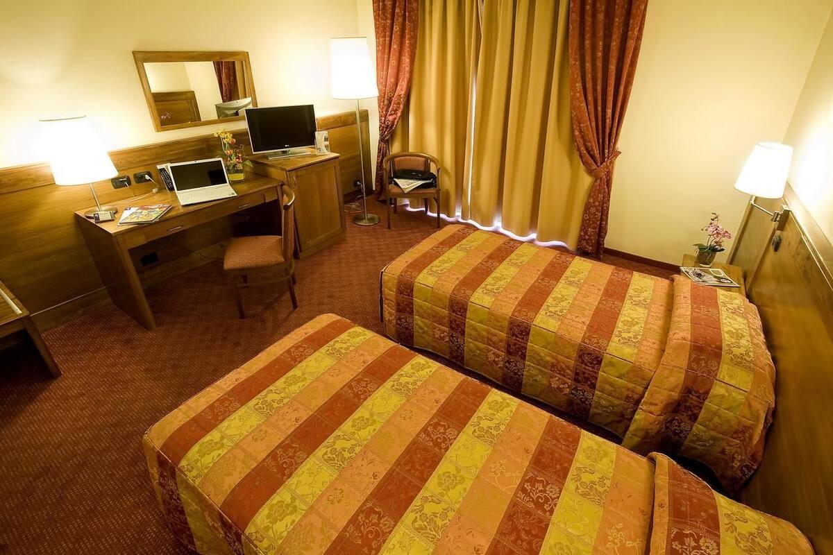 A hotel room with two beds, a desk with a laptop, a chair, and a small table with a lamp.