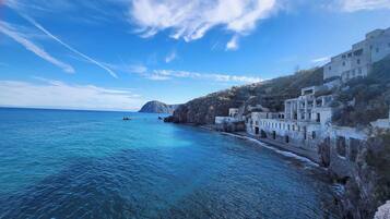 Paesaggio costiero con mare cristallino, scogliere rocciose e un edificio bianco con una scala che scende verso l'acqua.