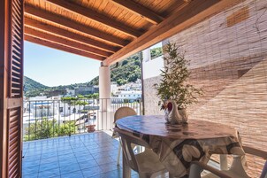 Un patio techado al aire libre con una mesa redonda y sillas, una planta en maceta y vistas de edificios y colinas al fondo.