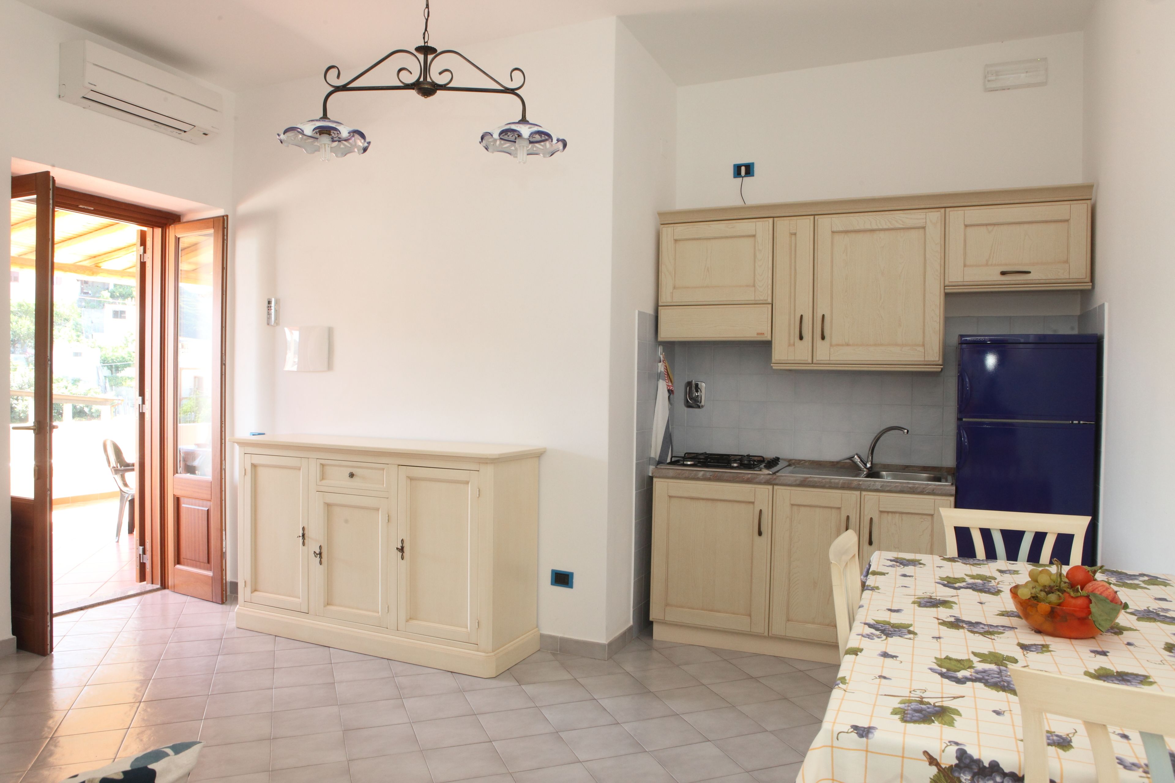 A kitchen with light wooden cabinets, a blue refrigerator, and a dining table with a fruit bowl.