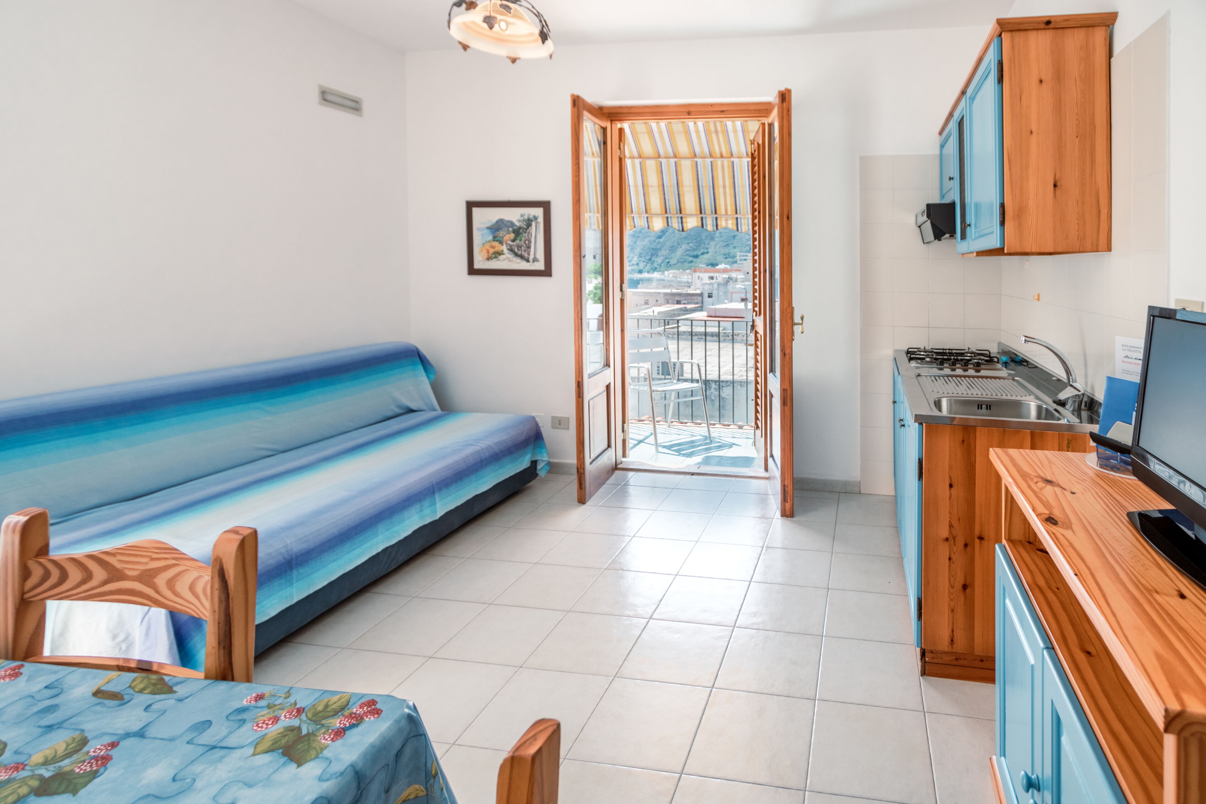 A room with a bed, a dining table, a kitchen area, and a view through an open door.