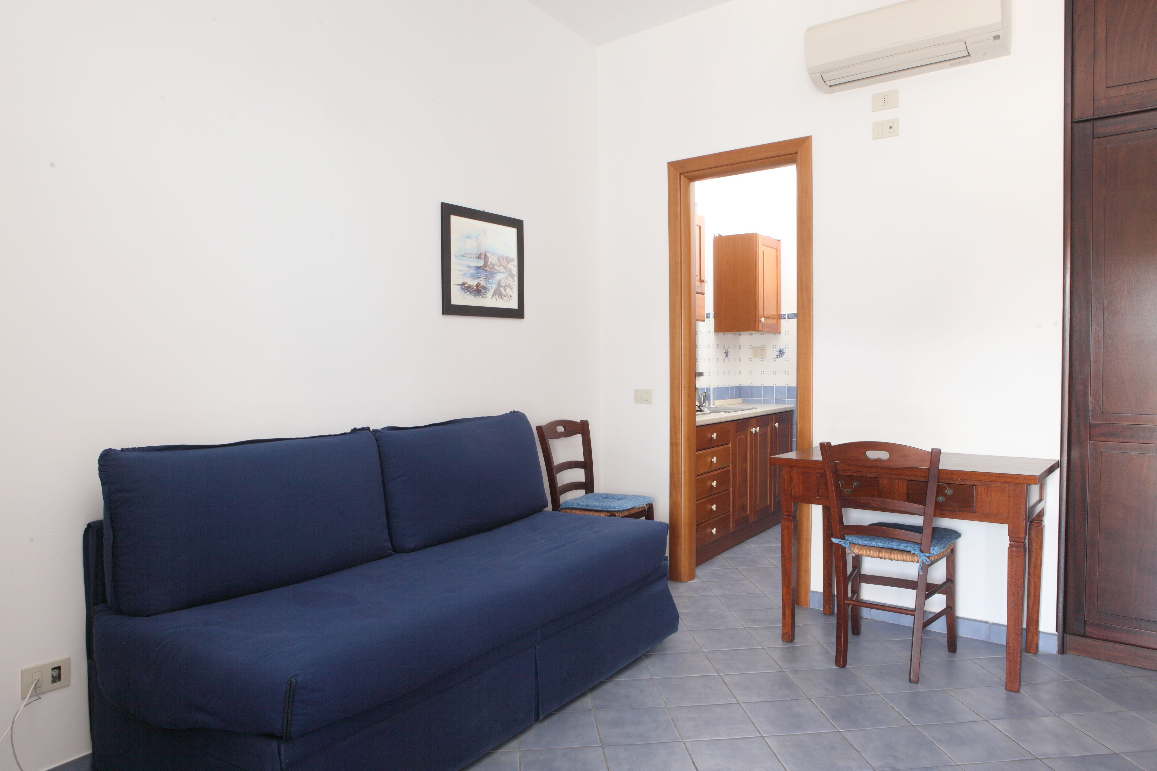A compact living space with a sofa, two chairs, a small table, and a kitchen area visible through an open door.