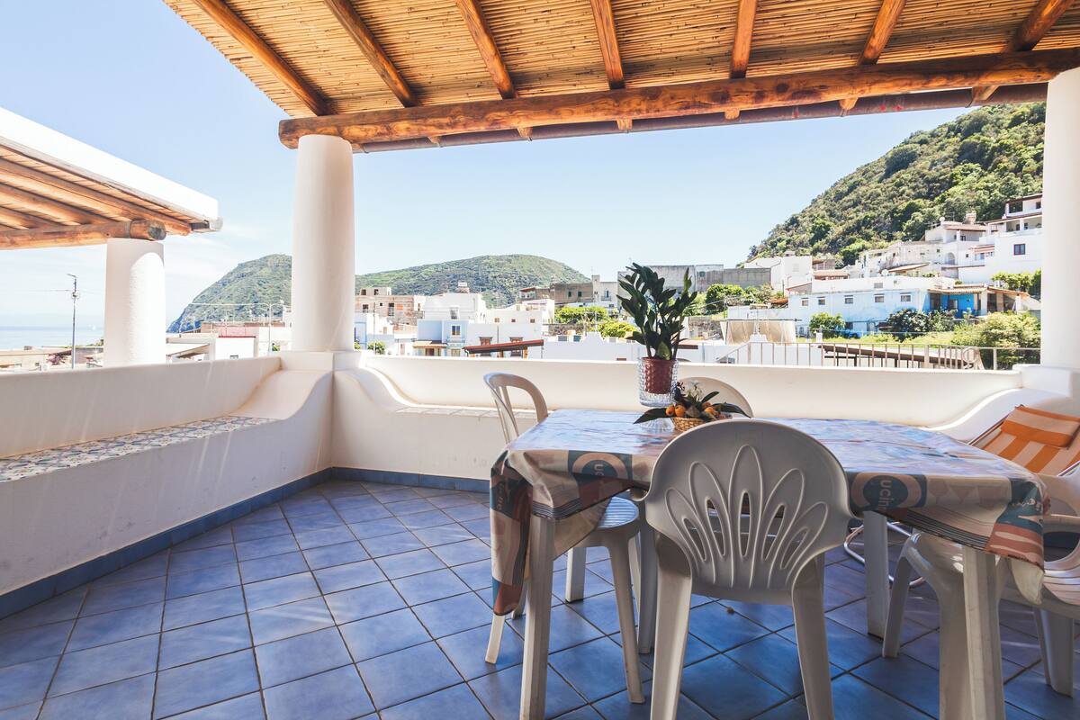 Un espace extérieur abrité, avec une table et des chaises, offrant une vue sur une ville perchée sur une colline, avec ses bâtiments et sa végétation.