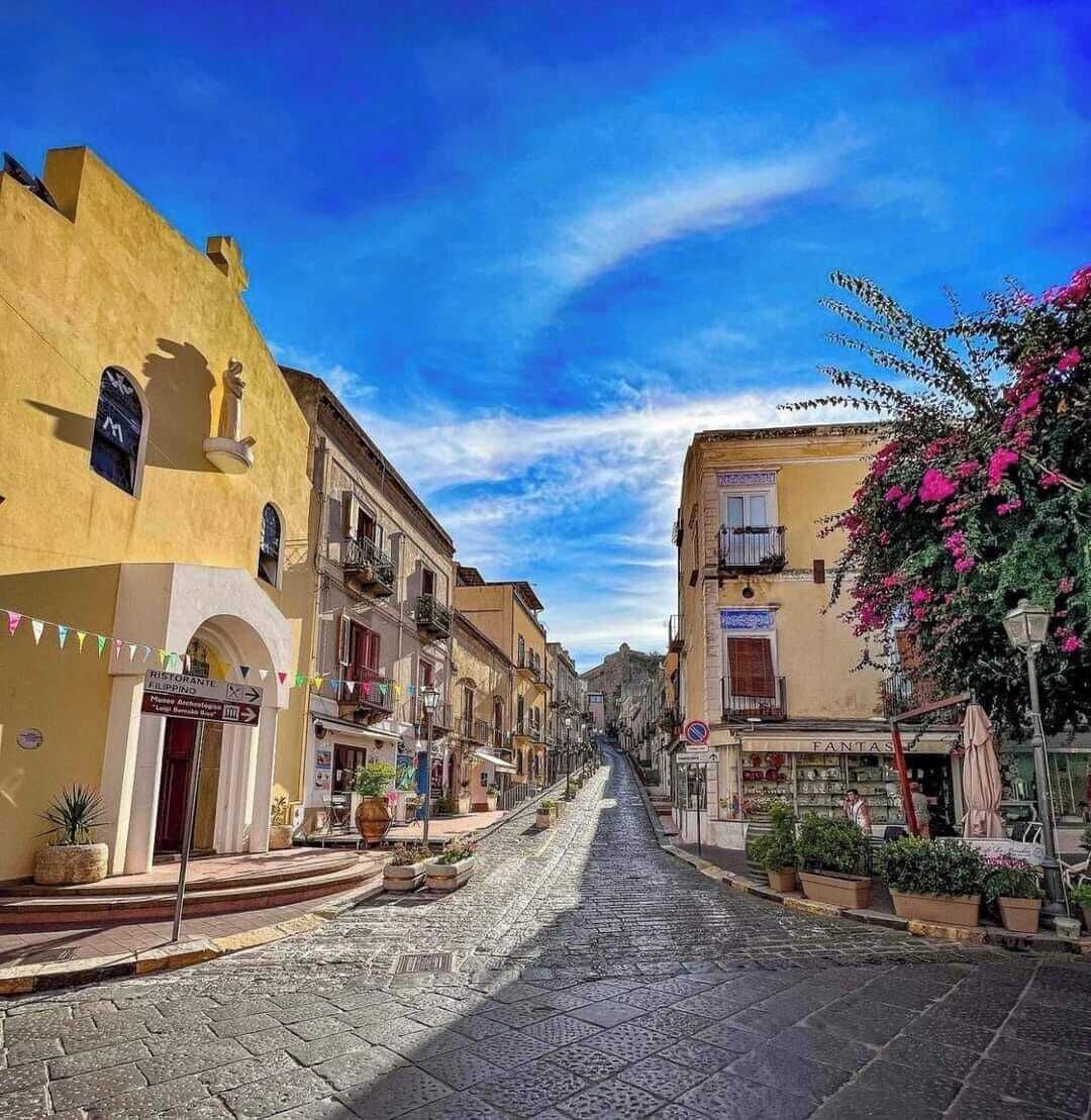 Una calle empedrada flanqueada por edificios históricos, un letrero de restaurante y macetas con plantas.