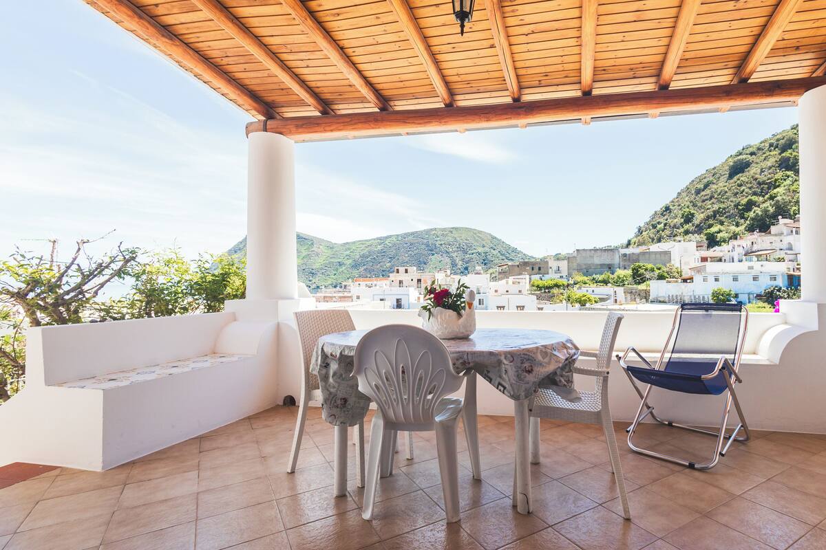 Une terrasse dotée d’un plafond en bois, de murs blancs et offrant une vue sur une ville perchée sur une colline.
