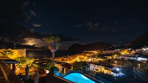 A nighttime view of a coastal town with a swimming pool, outdoor seating, and a view of the sea.