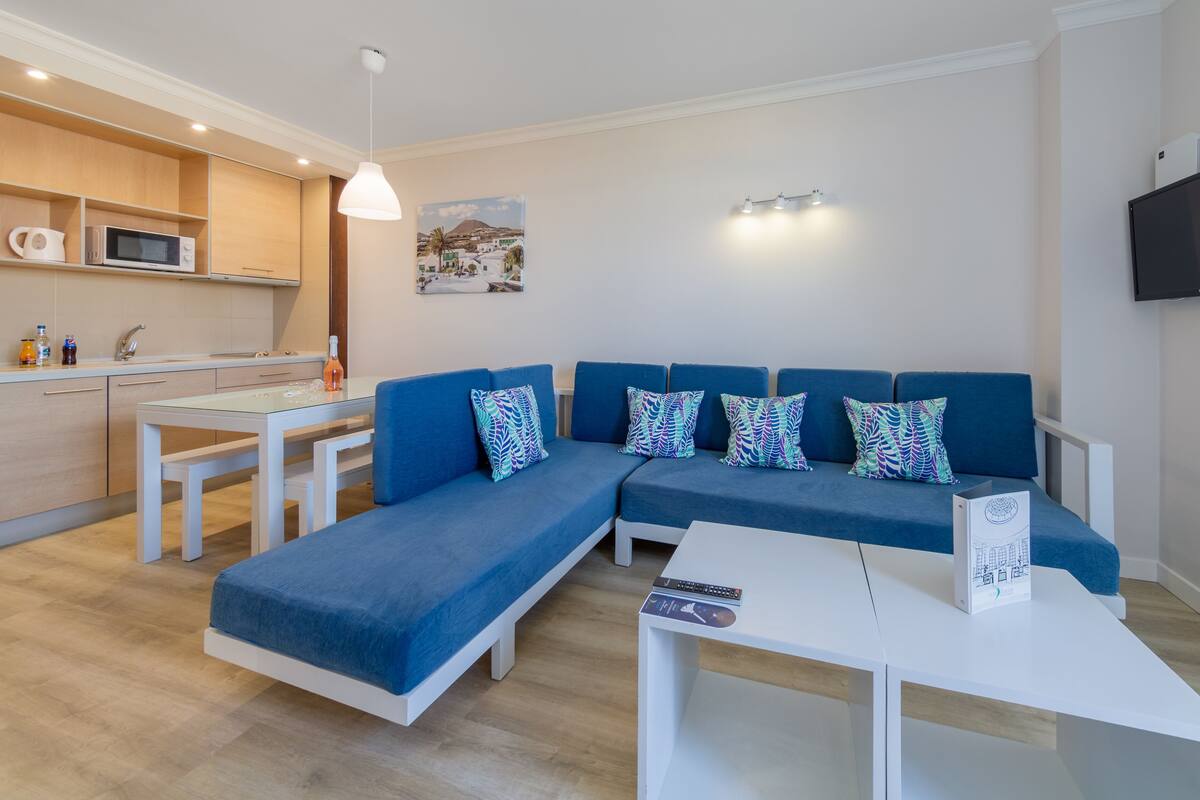 A modern living room with a blue sectional sofa, a white coffee table, and a kitchen area with a microwave and kettle.