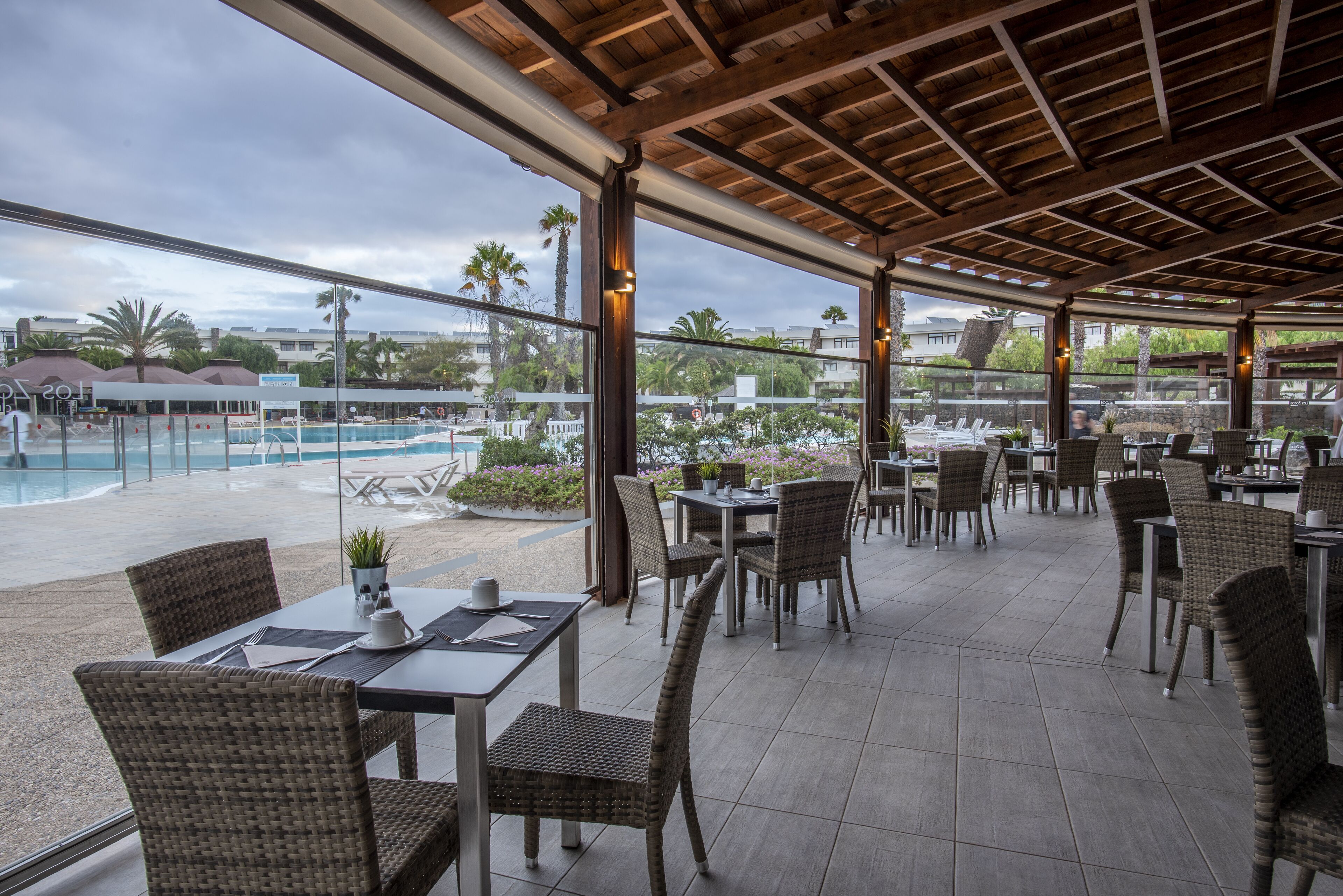 Un espace de restauration en plein air, avec des chaises et des tables en osier, donnant sur une piscine et des palmiers.