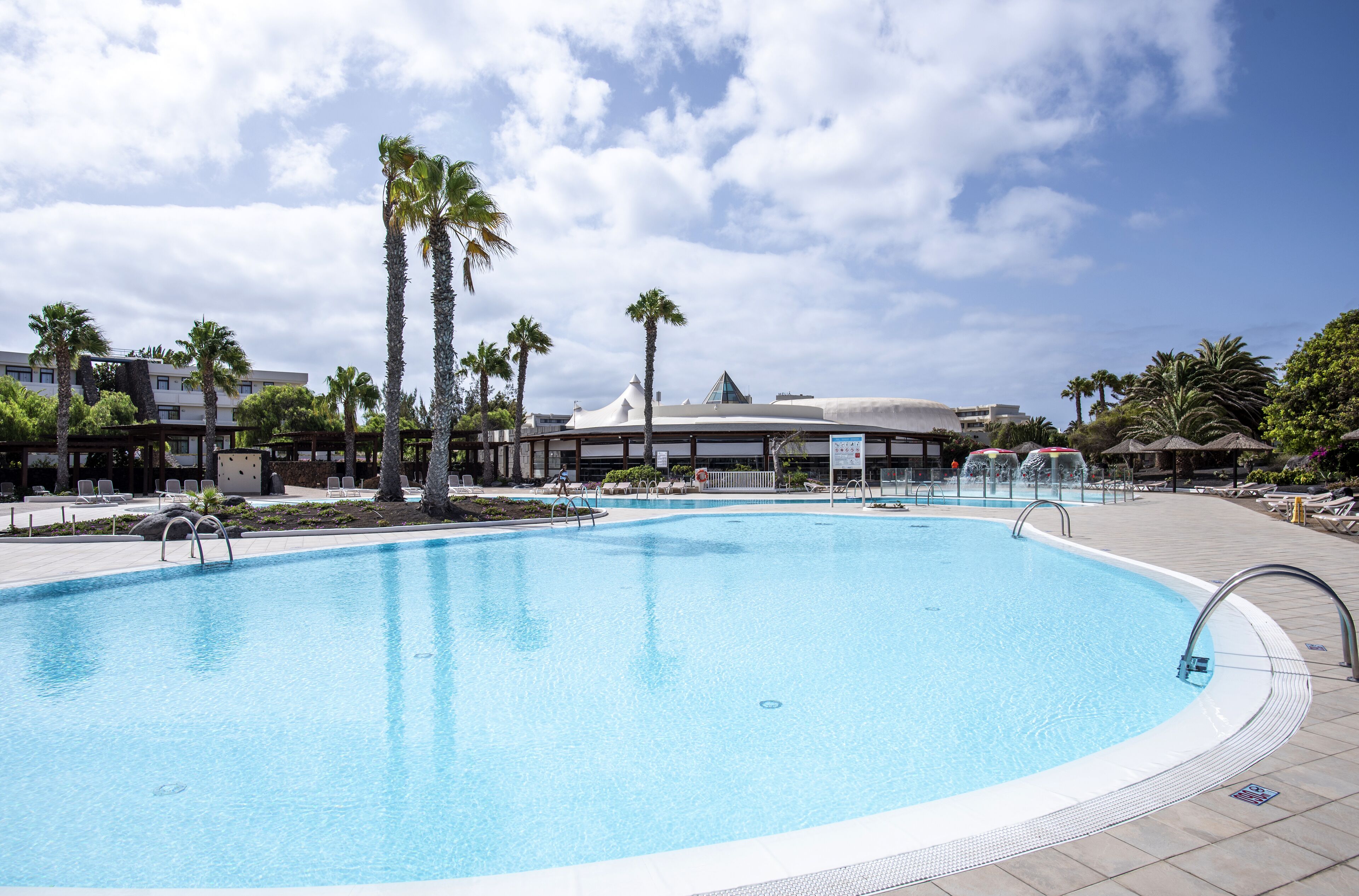 Una piscina con palmeras, sillones de descanso y un edificio al fondo.