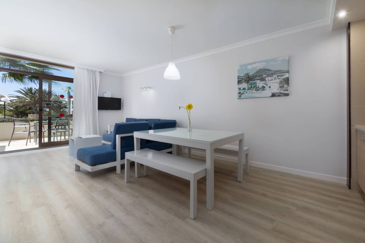 A modern living room with a glass dining table, blue sofas, a white coffee table, and a painting on the wall.