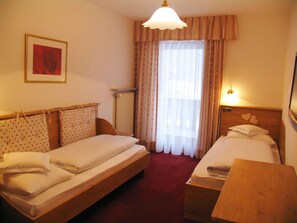 Une chambre d’hôtel avec deux lits simples, une tête de lit en bois, un radiateur, une fenêtre avec des rideaux, un luminaire fixé au mur et un tableau encadré.