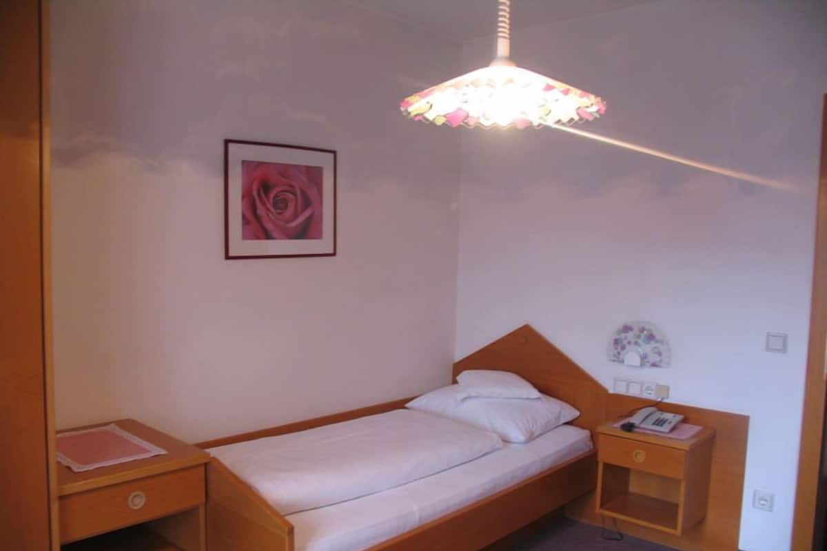 A single bed with a wooden headboard, a bedside table with a lamp, a small shelf, and a framed picture on the wall.