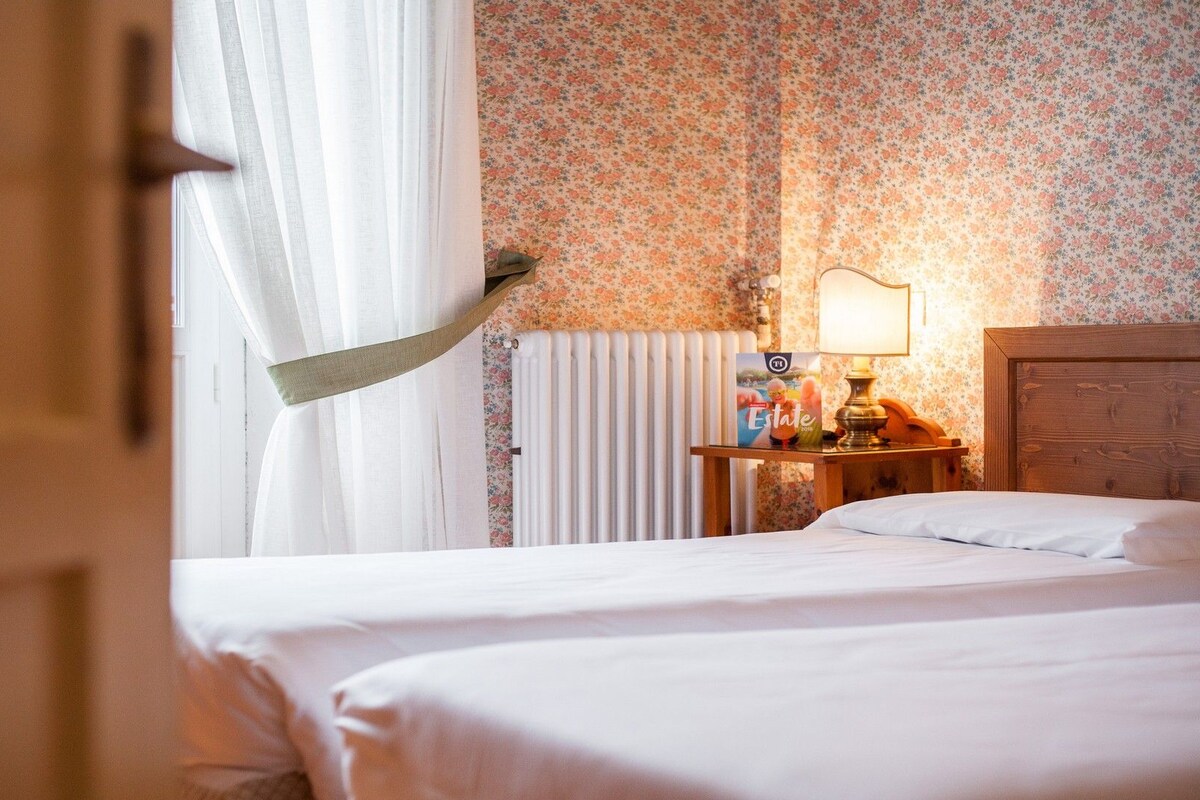 Une chambre d’hôtel avec un lit, une table de chevet avec une lampe, un papier peint à motifs floraux et une fenêtre avec des rideaux.