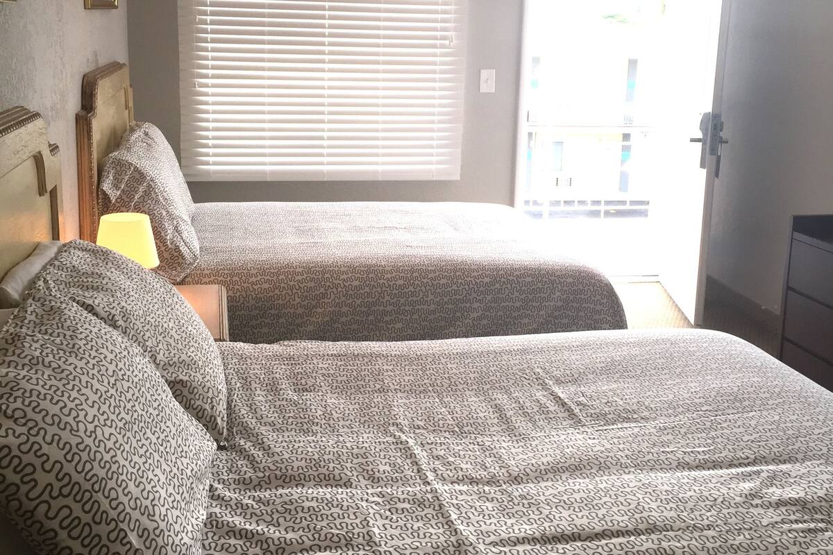 A bedroom with two beds, a window with blinds, a bedside lamp, and a door leading outside.
