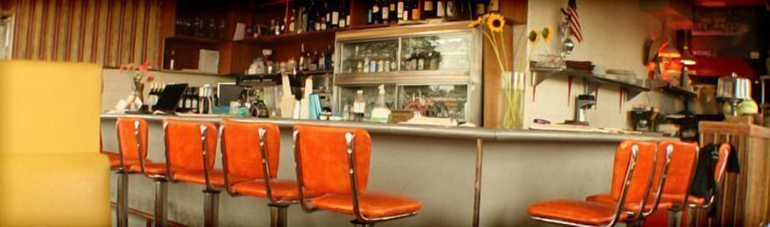 A bar with orange bar stools, a counter with a cash register, and shelves with bottles and glasses.