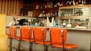 En bar med orange barstole, en disk med kasseapparat og hylder med flasker og glas.