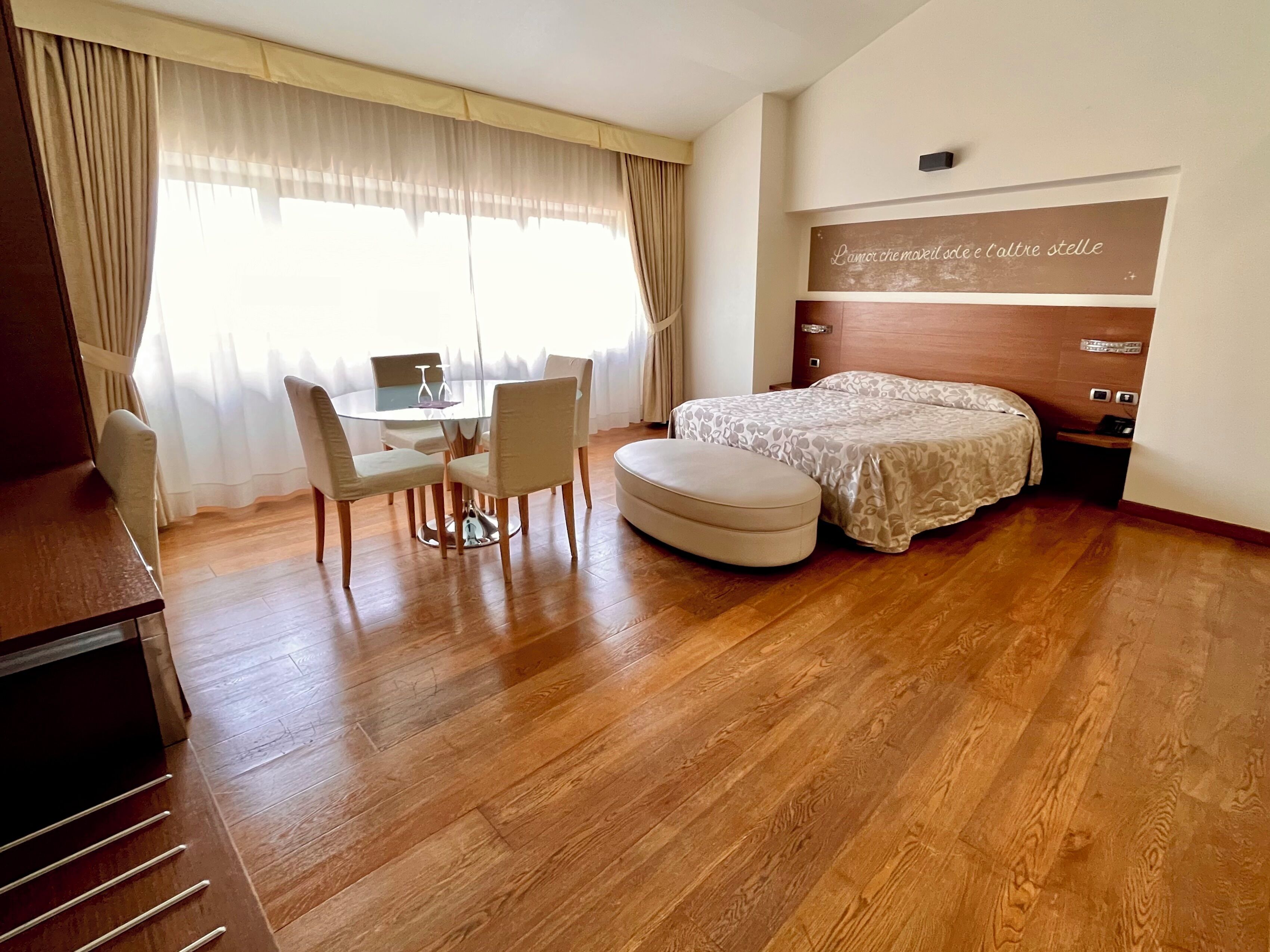 A hotel room with a wooden floor, a bed with a patterned bedspread, a small table with chairs, and a desk with a computer.