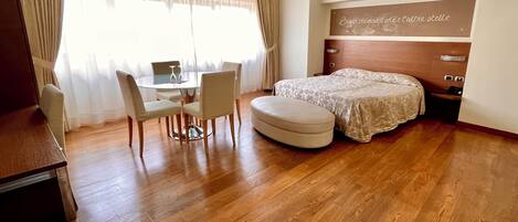 Habitación de hotel con piso de madera, una cama con colcha estampada, una mesita con sillas y un escritorio con computadora.
