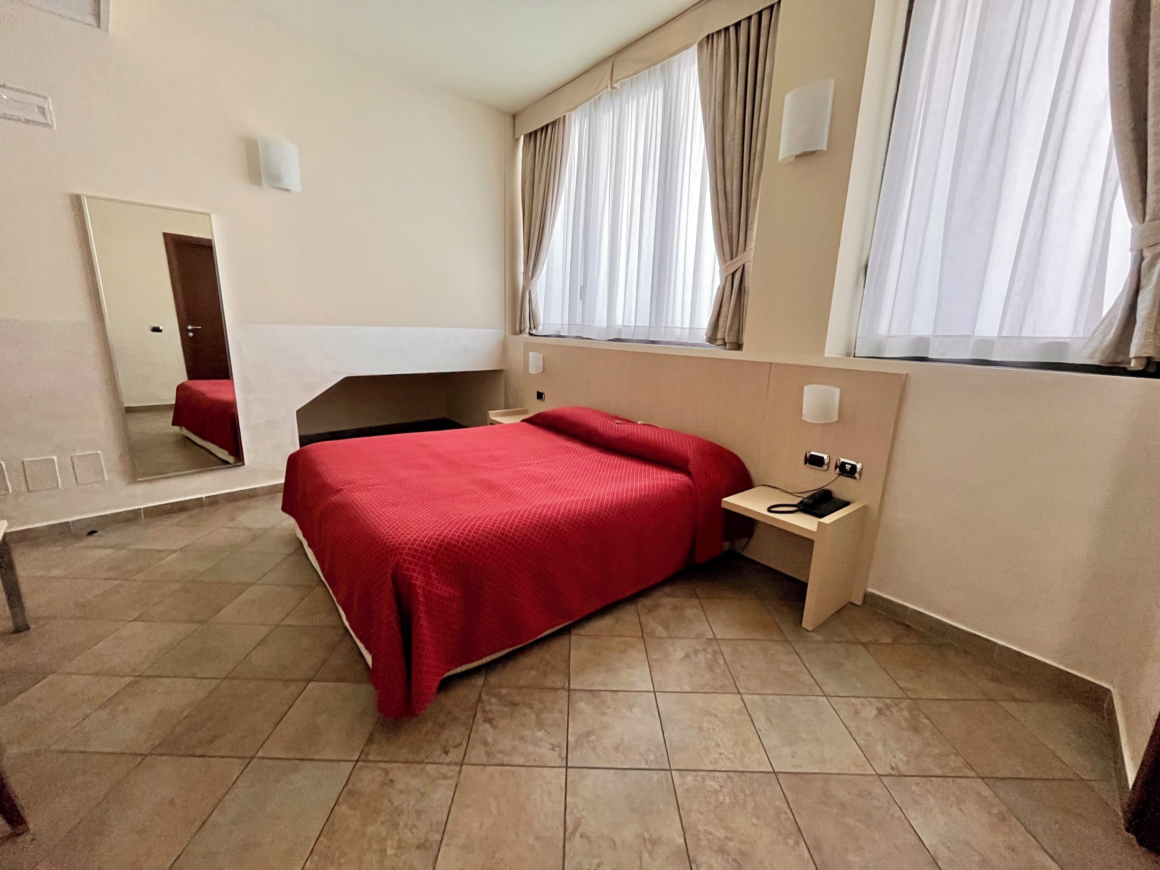 A hotel room with a bed, a bedside table, a mirror, and a window with curtains.