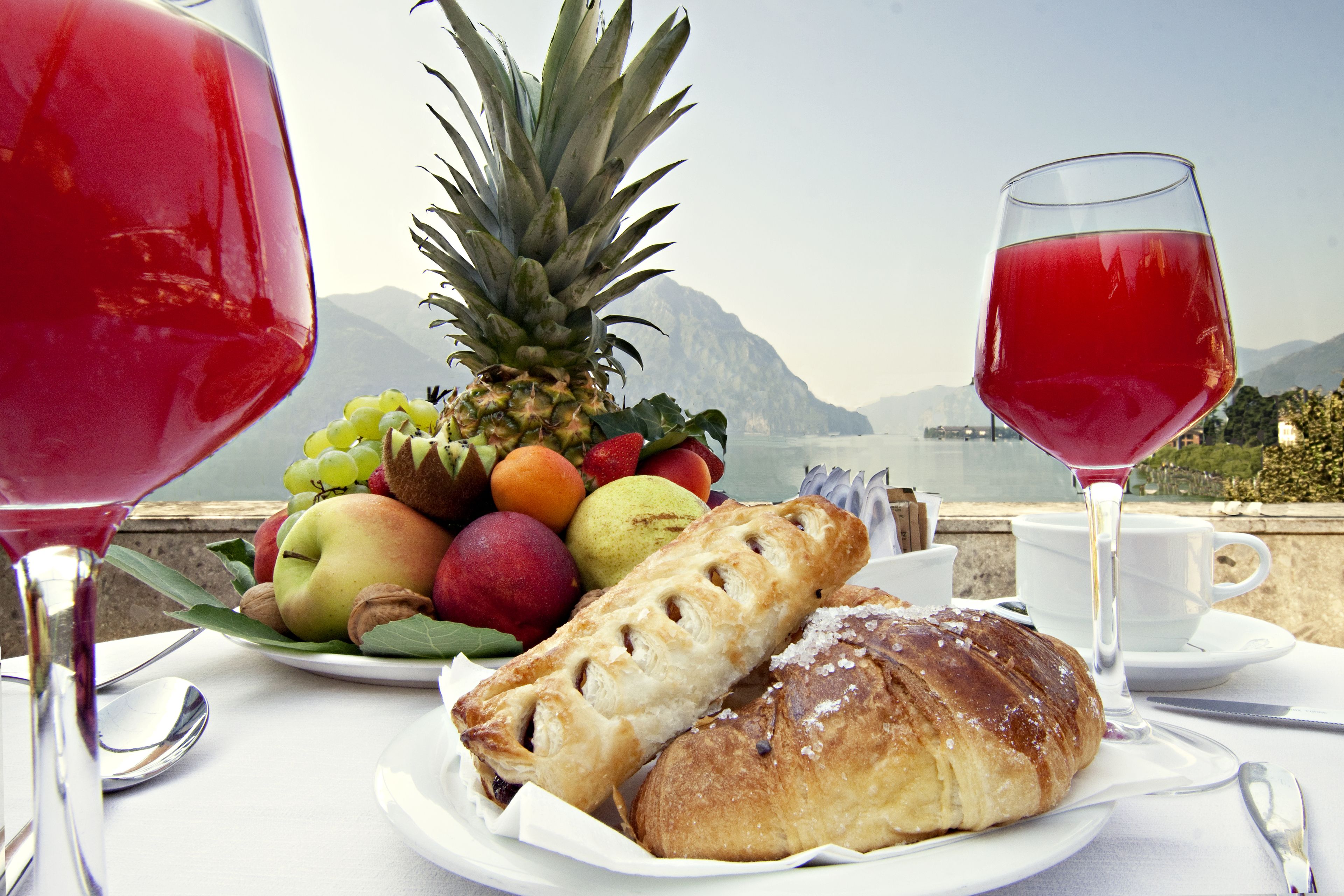 A table with a croissant, a pastry, and a fruit plate, along with two glasses of red wine, set against a scenic mountain backdrop.