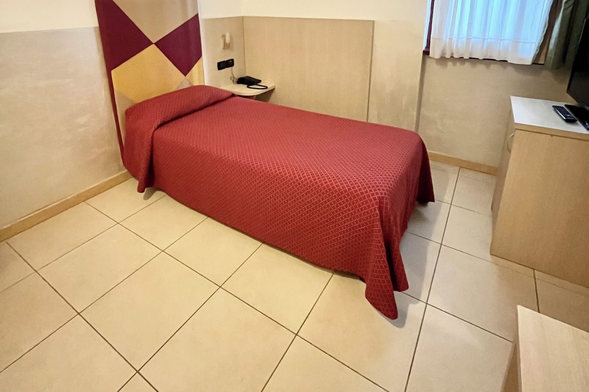 Ein Hotelzimmer mit einem Einzelbett, einem Schreibtisch und einem Fenster mit Vorhängen.