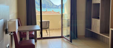A hotel room with a bed, a desk, a chair, a wardrobe, and a balcony with a view of the sea and mountains.