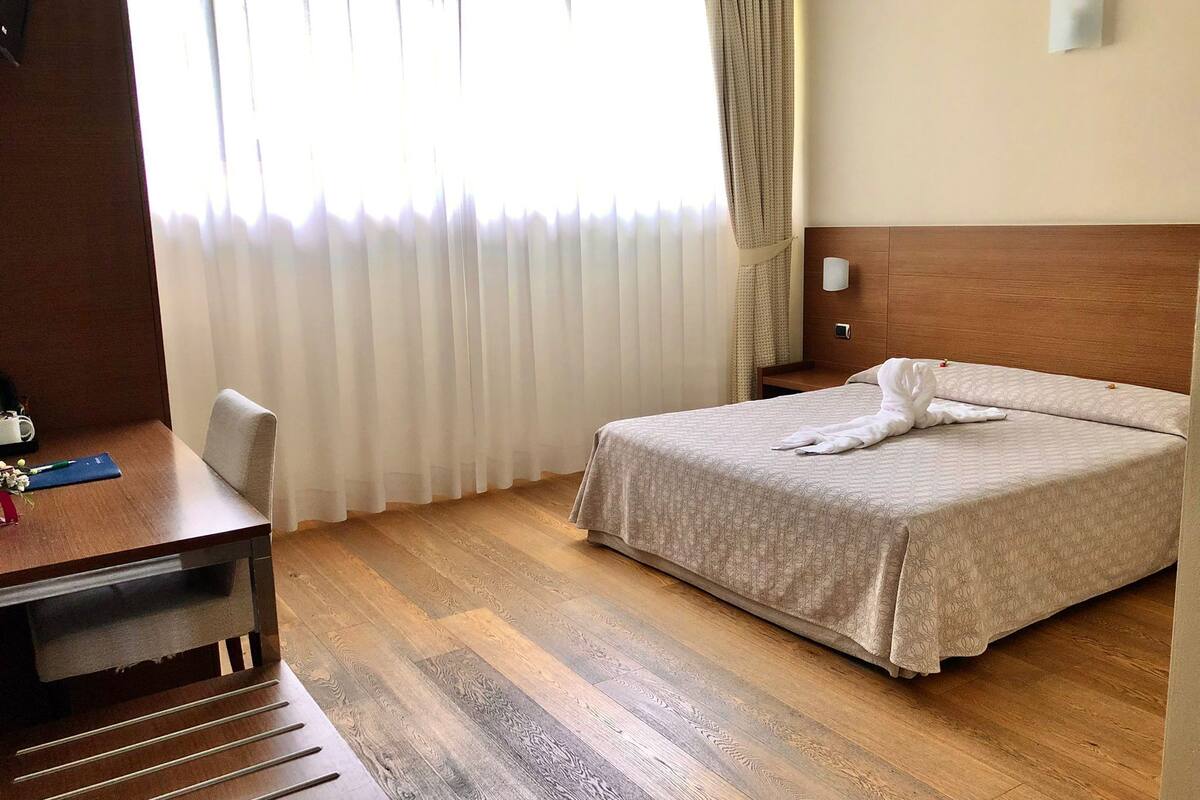 Ein Hotelzimmer mit Holzboden, einem Bett mit gemustertem Überwurf, einem Schreibtisch mit Stuhl und einem Fenster mit durchscheinenden Vorhängen.
