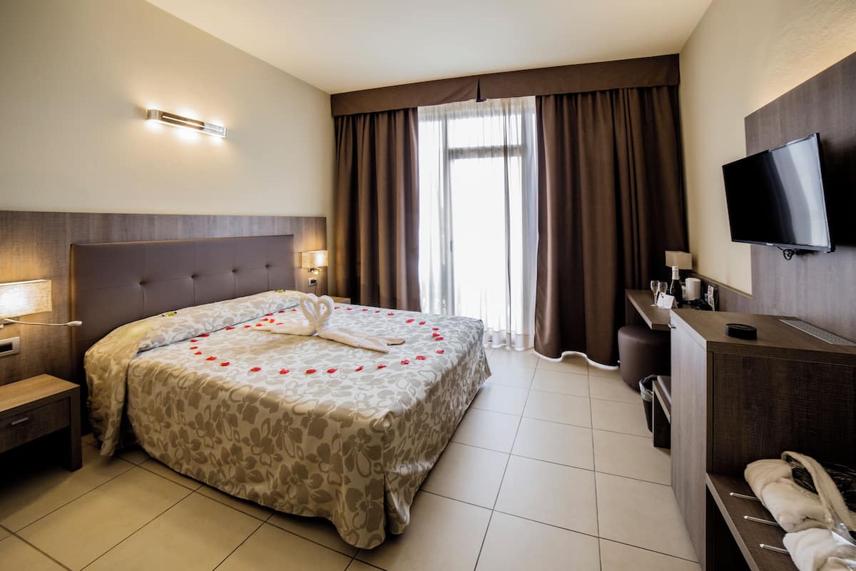 Ein Hotelzimmer mit einem großen Bett, einem an der Wand montierten Fernseher, einem Schreibtisch mit Lampe und einem Fenster mit Vorhängen.