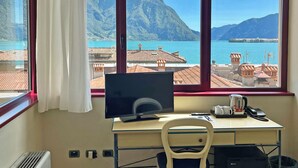 A room with a large window overlooking a mountainous landscape and a body of water. A desk with a computer, chair, and a small black refrigerator is visible. - Hotel Lovere Resort & Spa (Lovere)