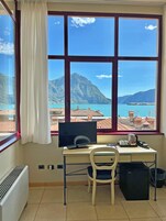 Una habitación con un gran ventanal que da a un paisaje montañoso y un cuerpo de agua. Se ve un escritorio con un ordenador, una silla y un refrigerador negro pequeño.