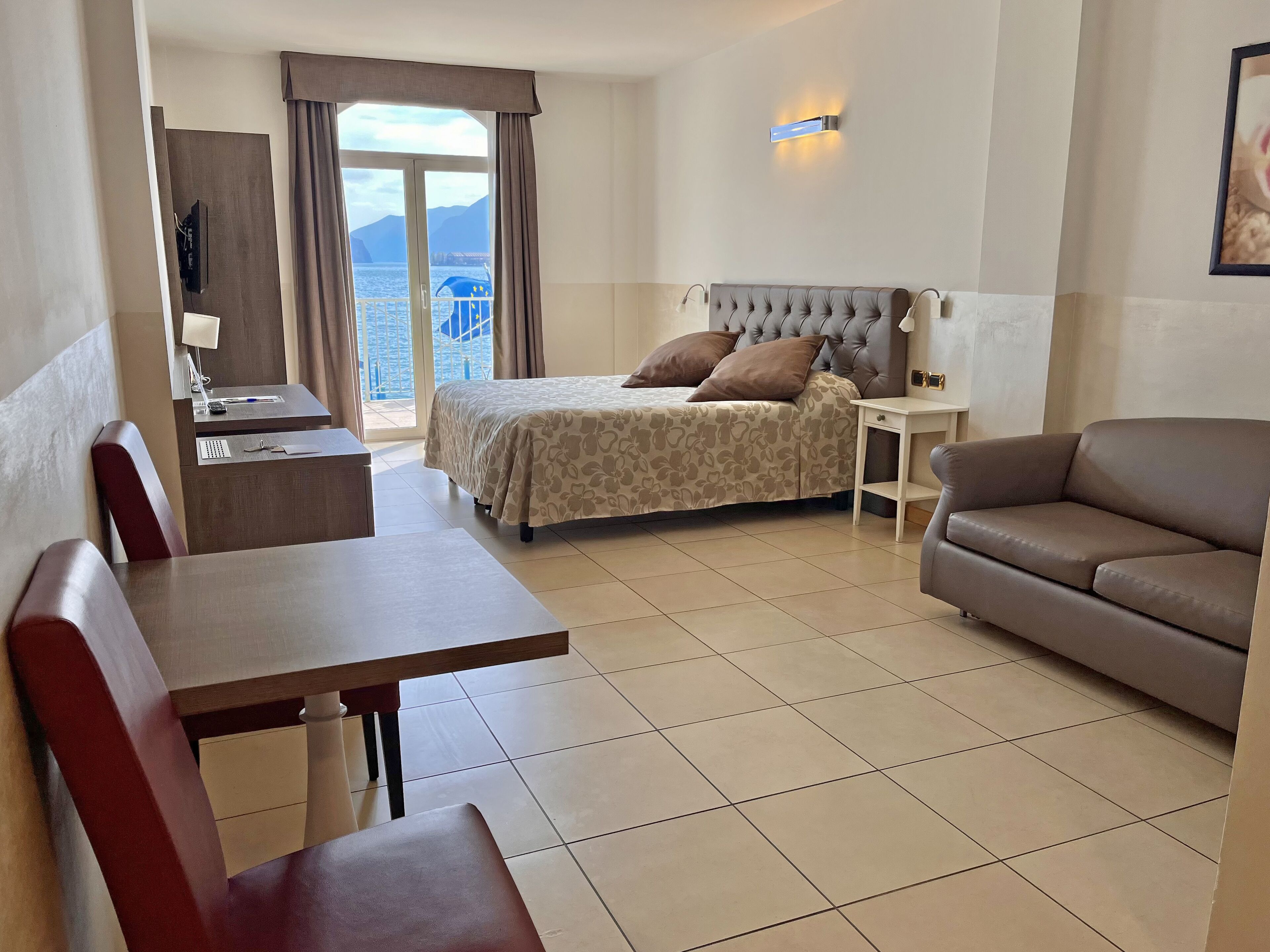 A hotel room with a bed, a desk, a chair, a sofa, and a view of the sea.