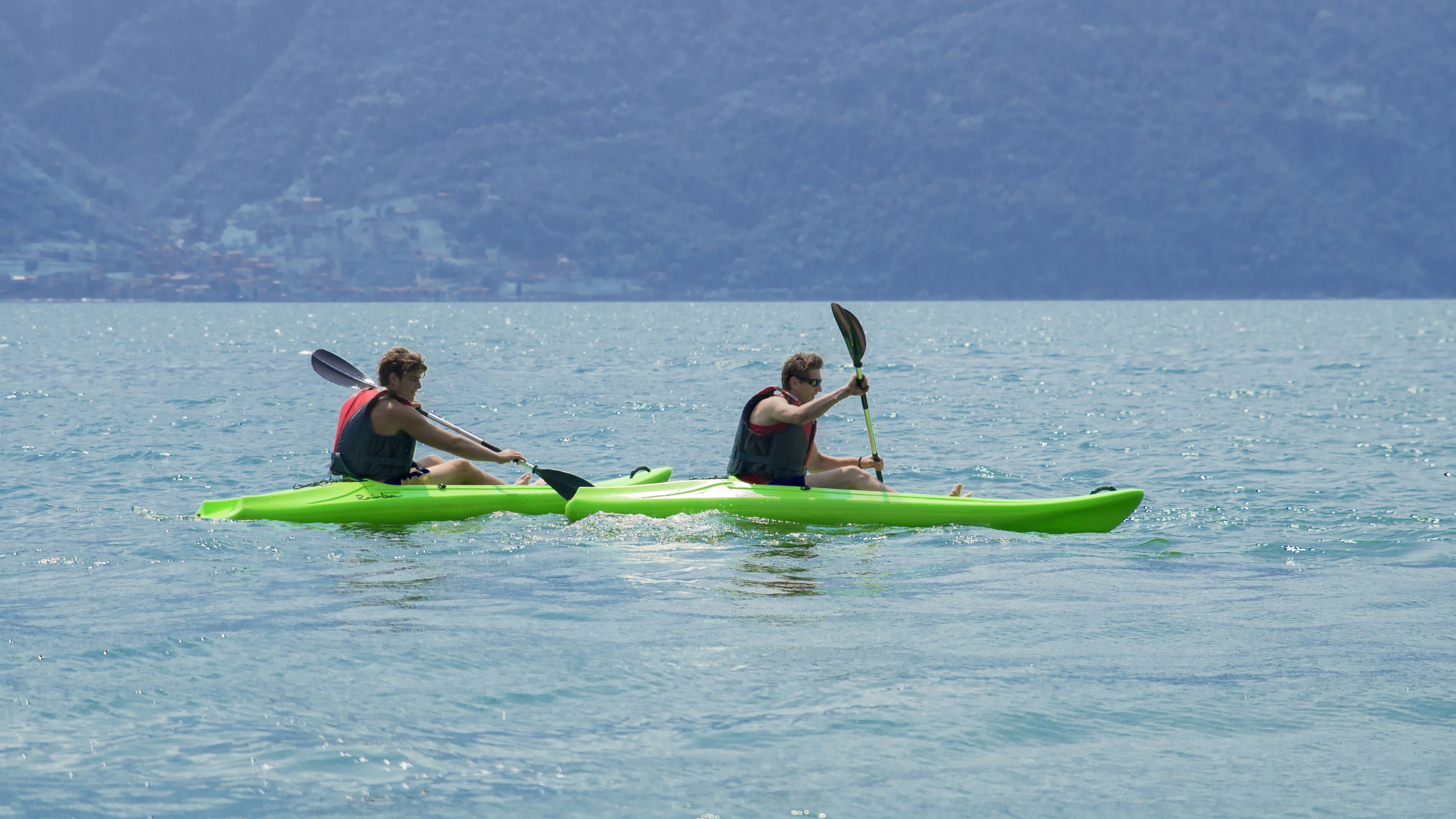 Two people kayaking on a calm body of water with a mountainous backdrop.