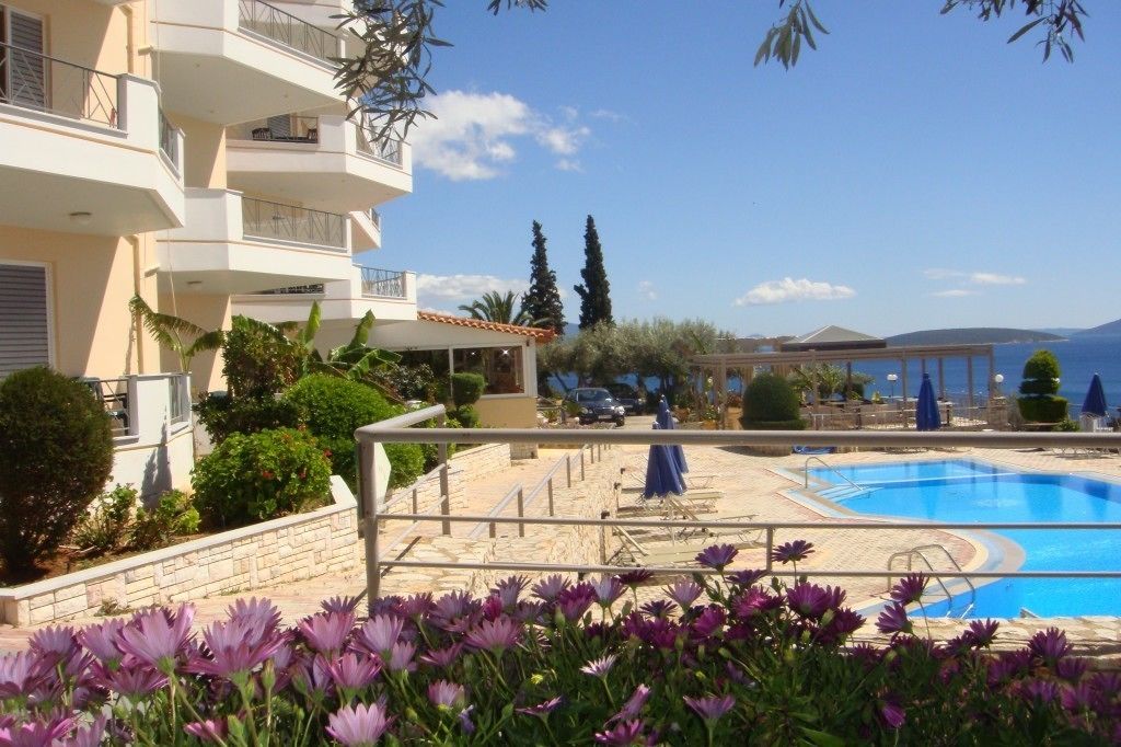Un espace piscine offrant une vue dégagée sur la mer, entouré d’un bâtiment doté de balcons et d’un jardin agrémenté de fleurs violettes.