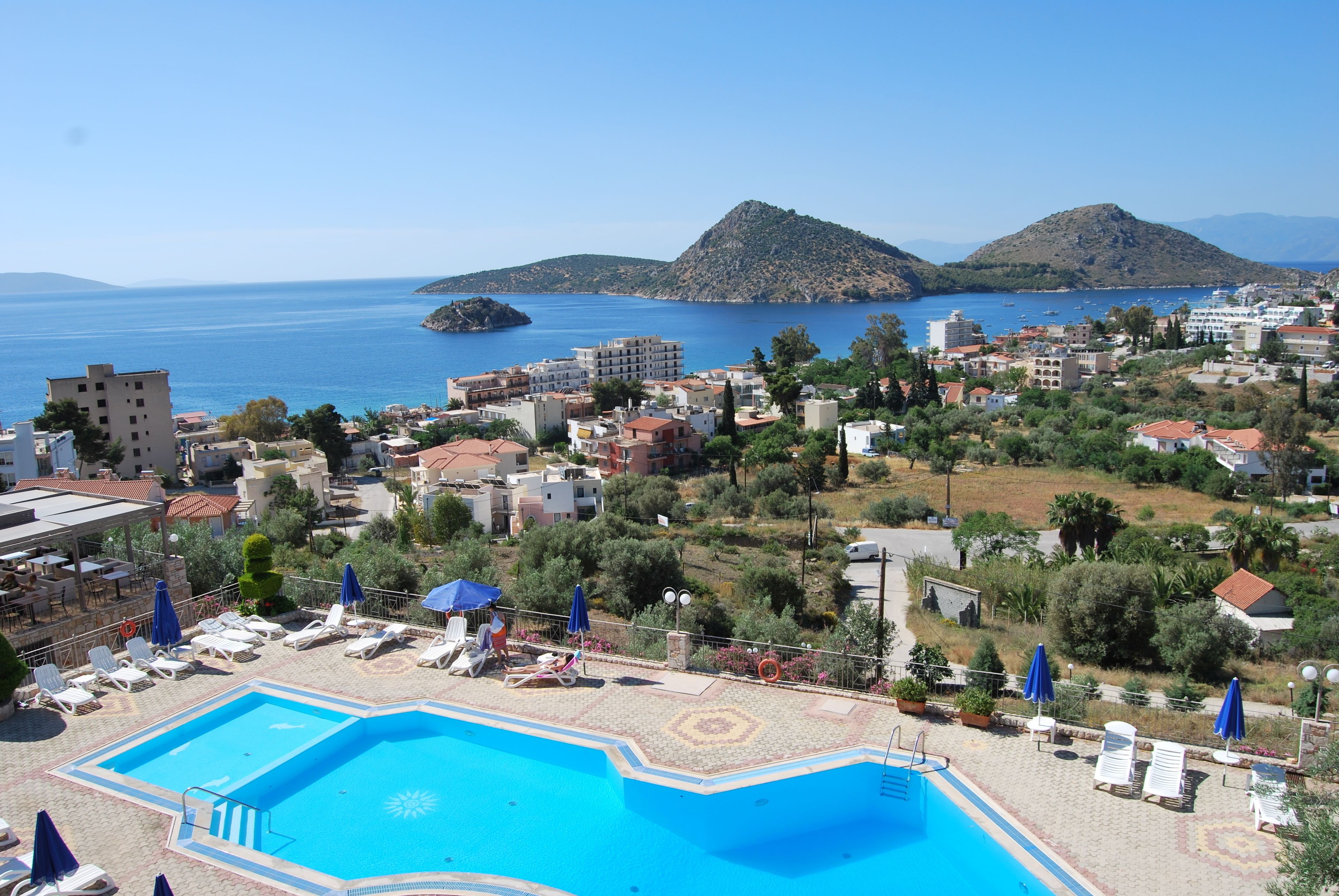 A swimming pool with lounge chairs and umbrellas, surrounded by buildings and a view of the sea and hills.