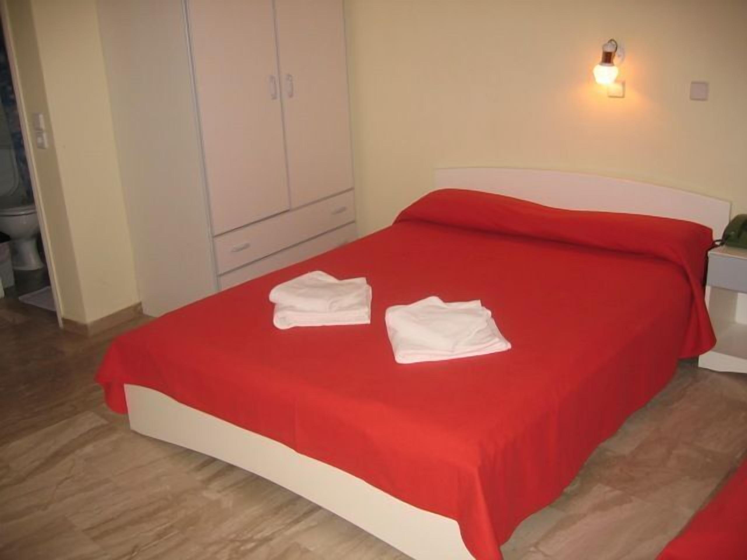 A bed with a red bedspread and two white towels, a white headboard, and a bedside table with a lamp.